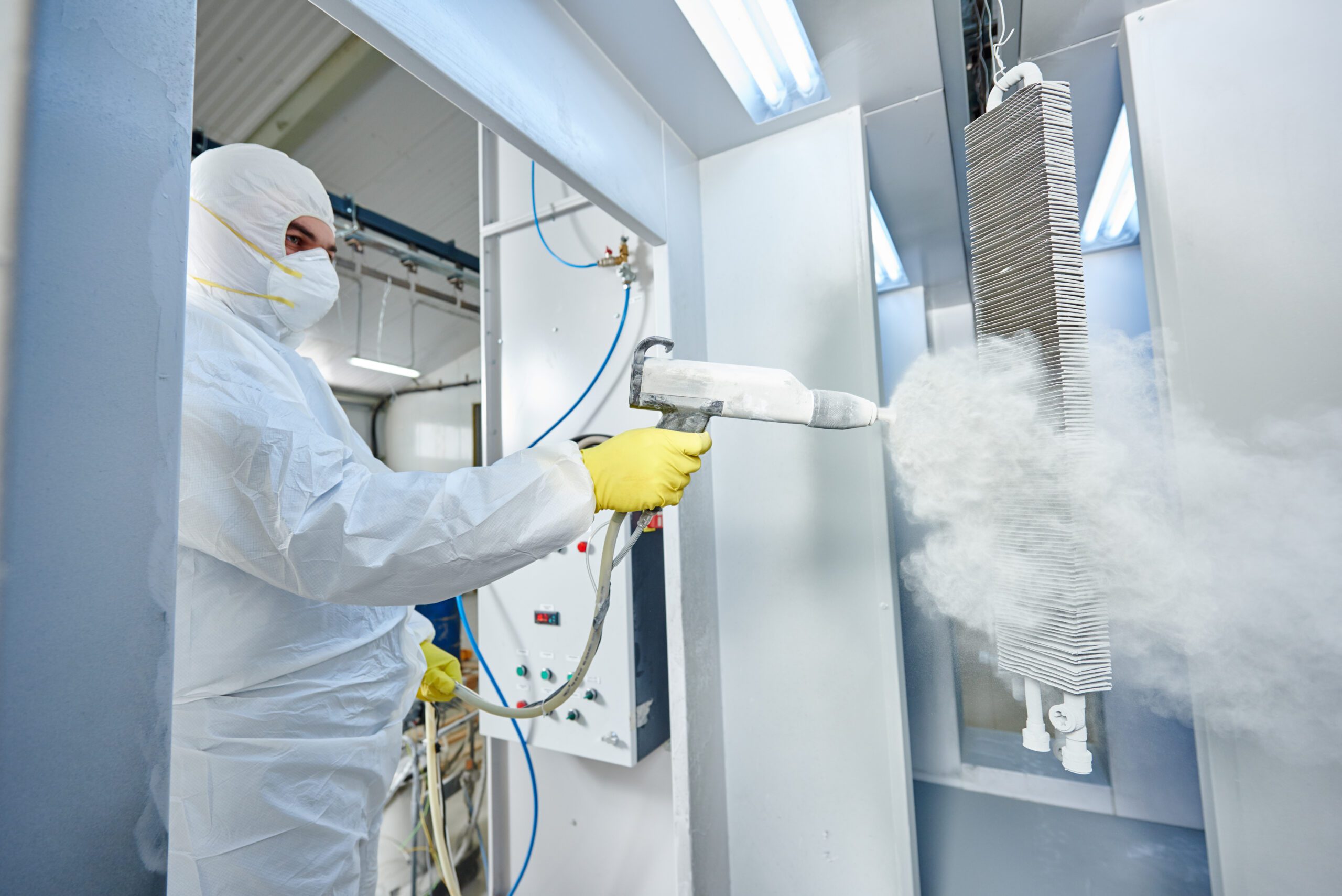 A man in a protective suit using a spray gun to apply powder coating to metal parts.