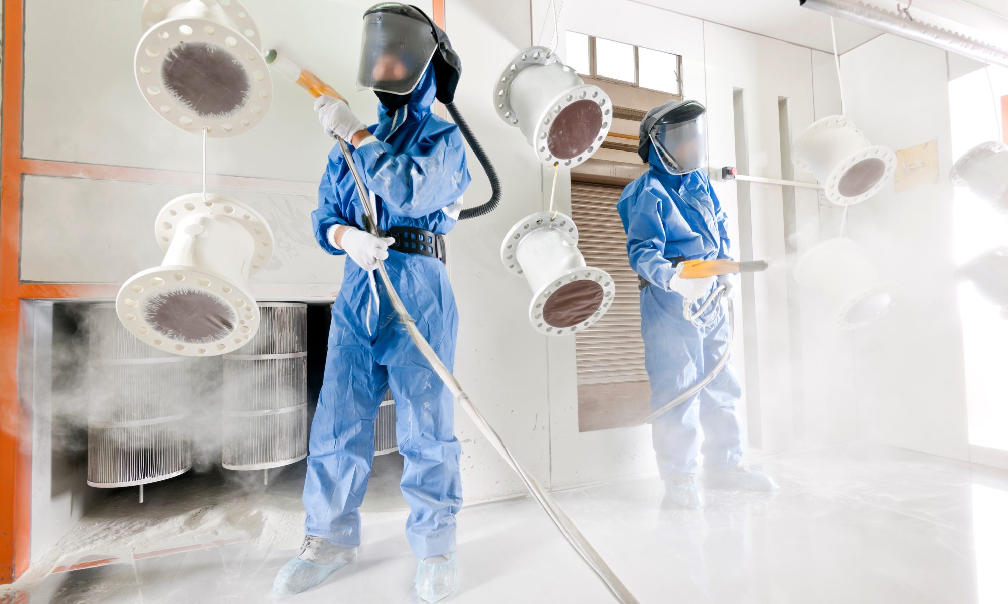 Two men in full paint suits and facemasks spray down tubing with powder-coating materials