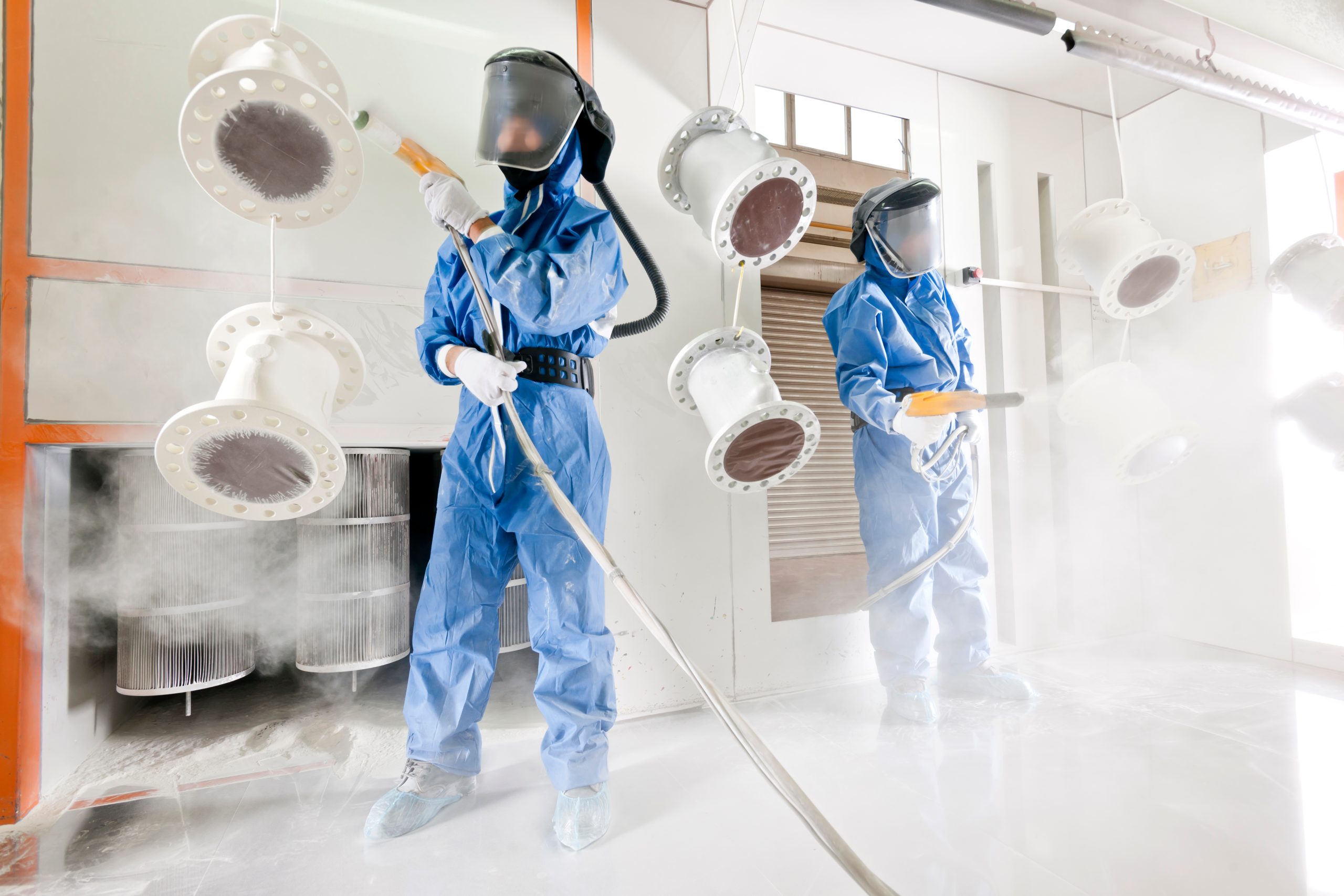 Two men in full paint suits and facemasks spray down tubing with powder-coating materials