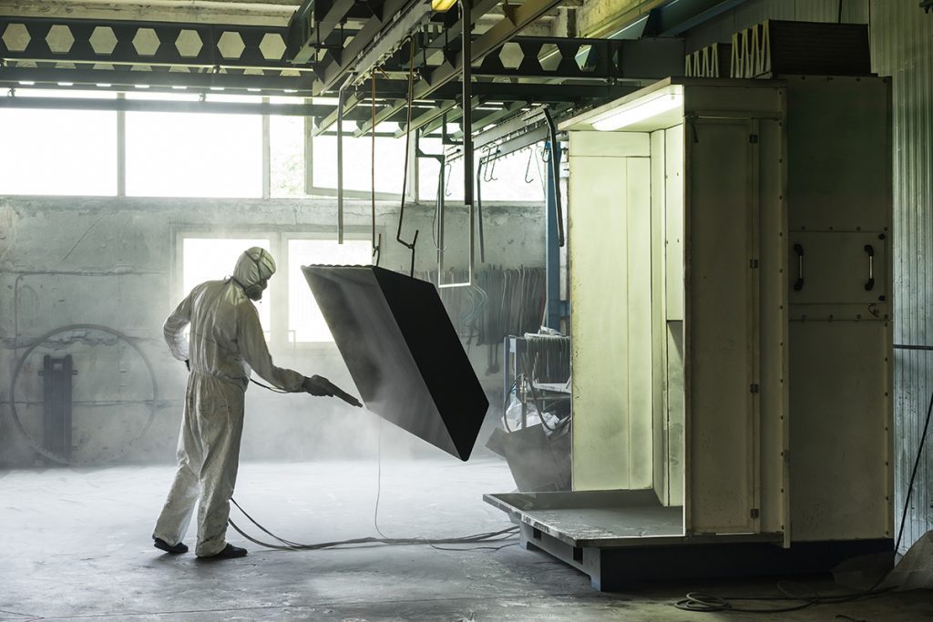 A worker in white PPE performs surface preparation in a warehouse by using sand-blasting gear