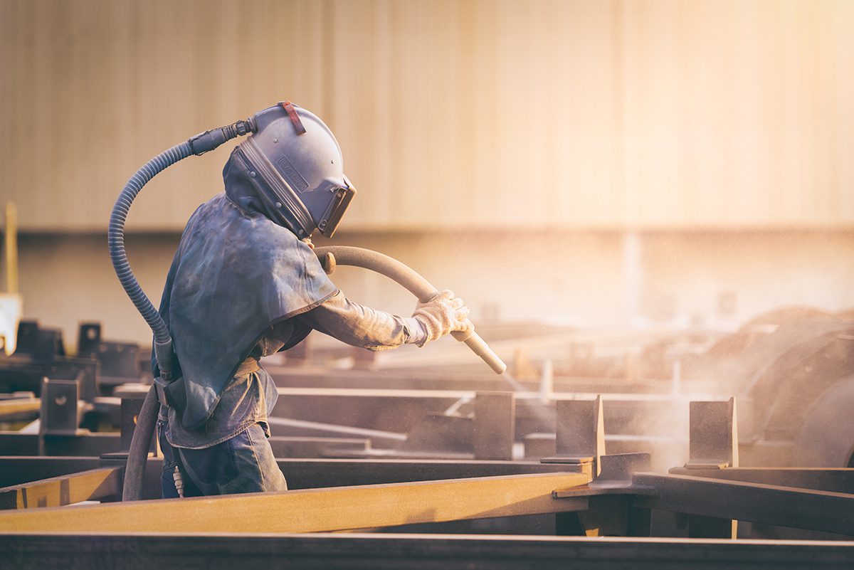 A worker in a blue full-body protective suit stands in the middle of large girders while wielding sand-blasting gear