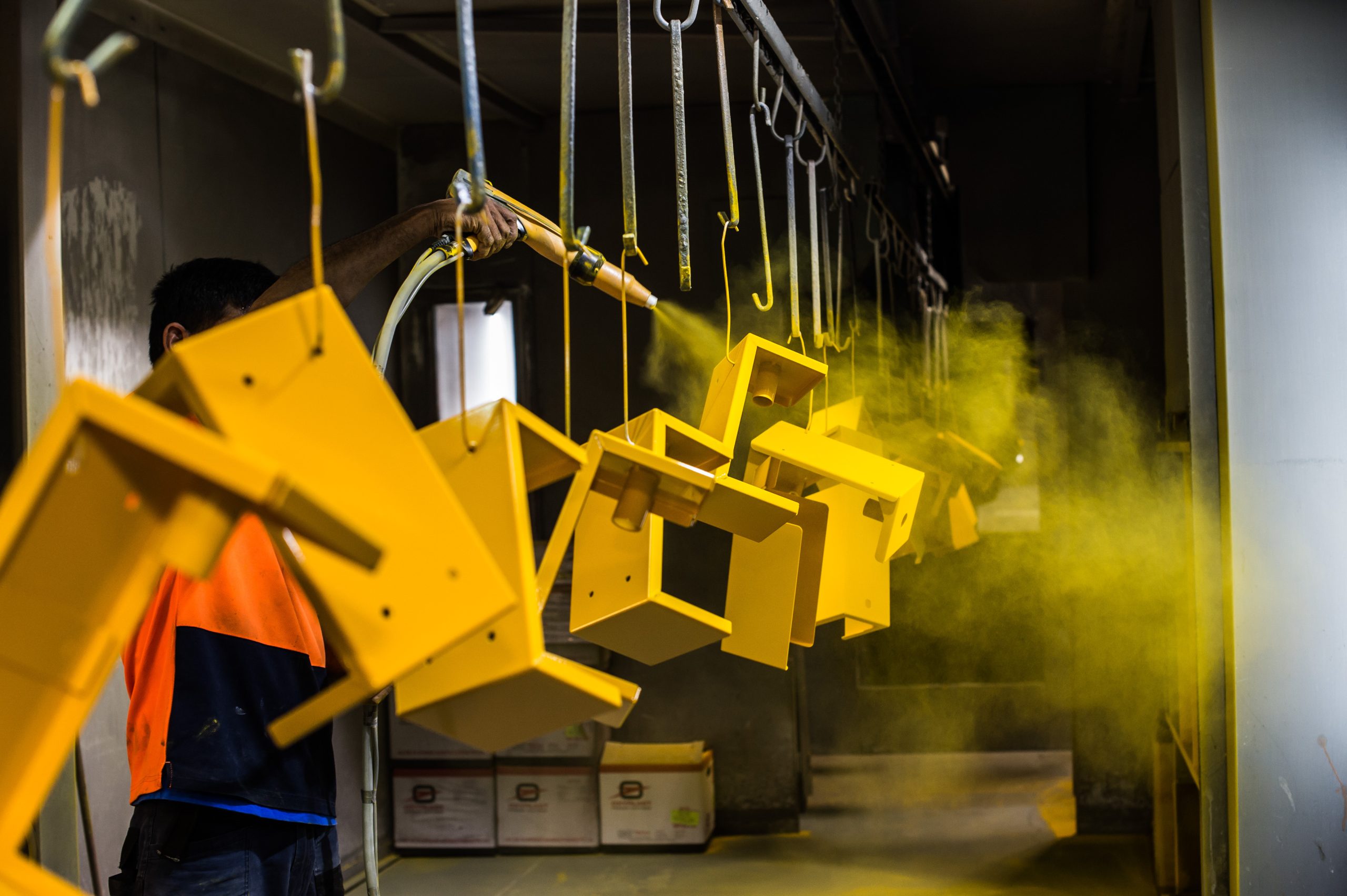 Pieces of a metal frame being sprayed with a bright yellow powder coating as they hang from a conveyor system.